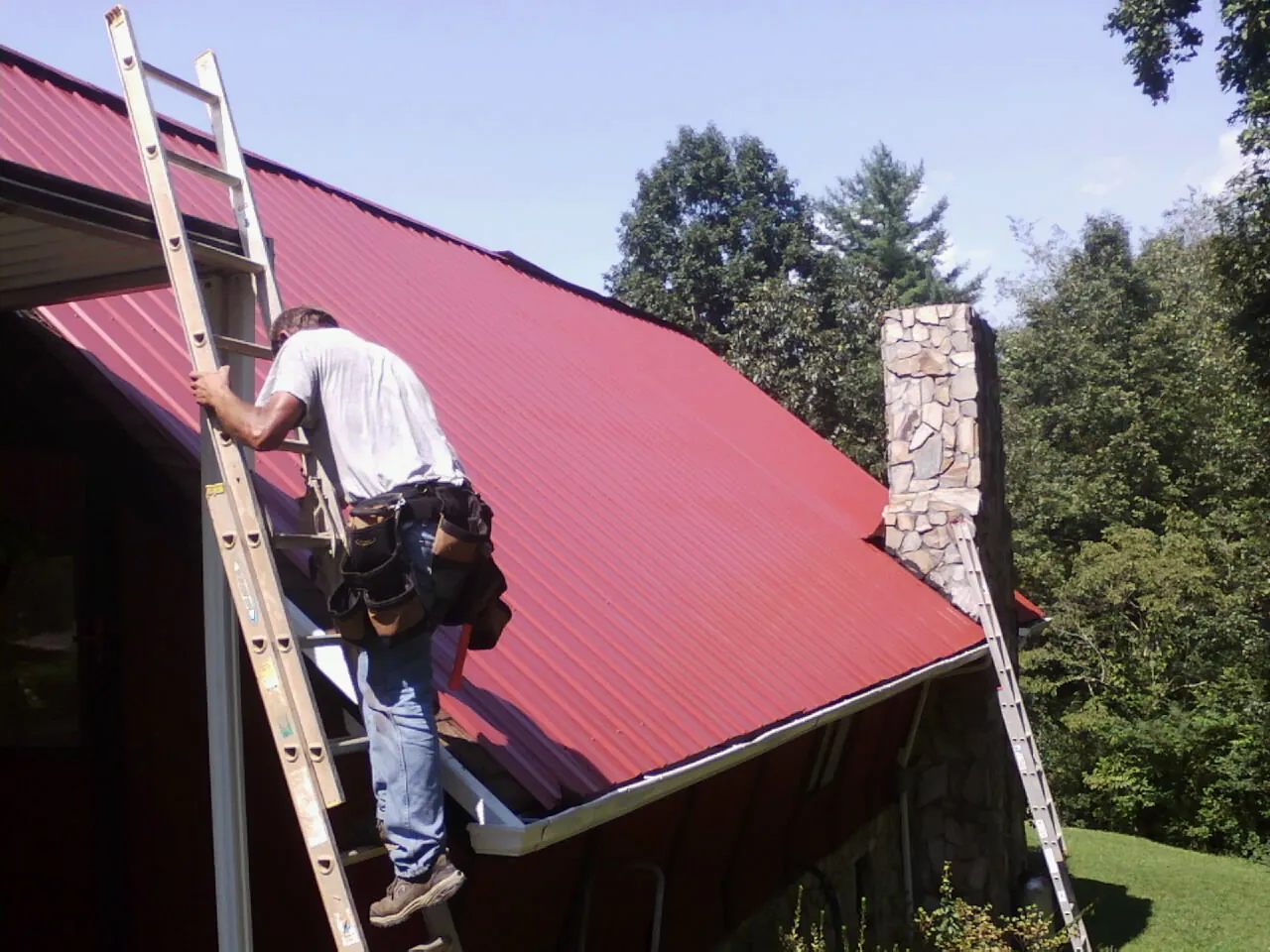 Roofer on a ladder beside a red metal roof and stone chimney — Soffit & Fascia Repair by Roofing Brothers in Somers